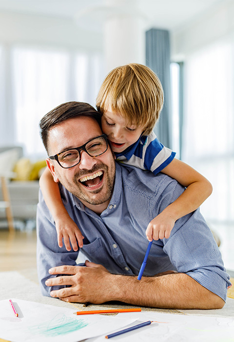 Un jeune père et son fils rient joyeusement dans le salon pour illustrer la page de l'assurance habitation Cocoon Flex de DVV